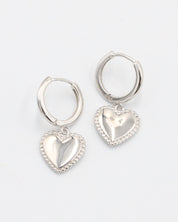 A pair of silver hoop earrings with heart-shaped pendants attached to them, displayed against a white background.