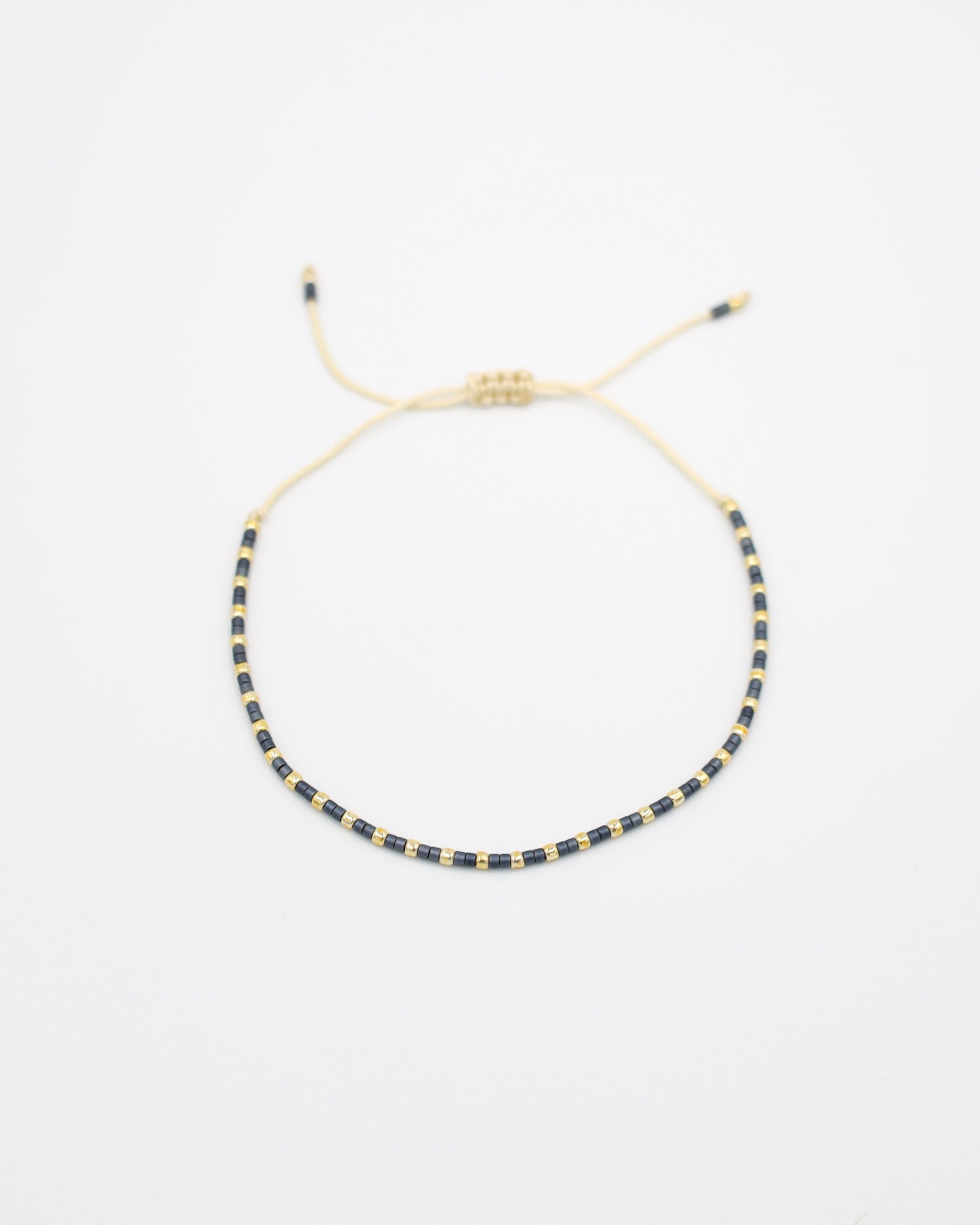 Flat-lay image of the Anya bracelet featuring alternating dark and gold beads, strung on a cream cord with an adjustable sliding closure.
