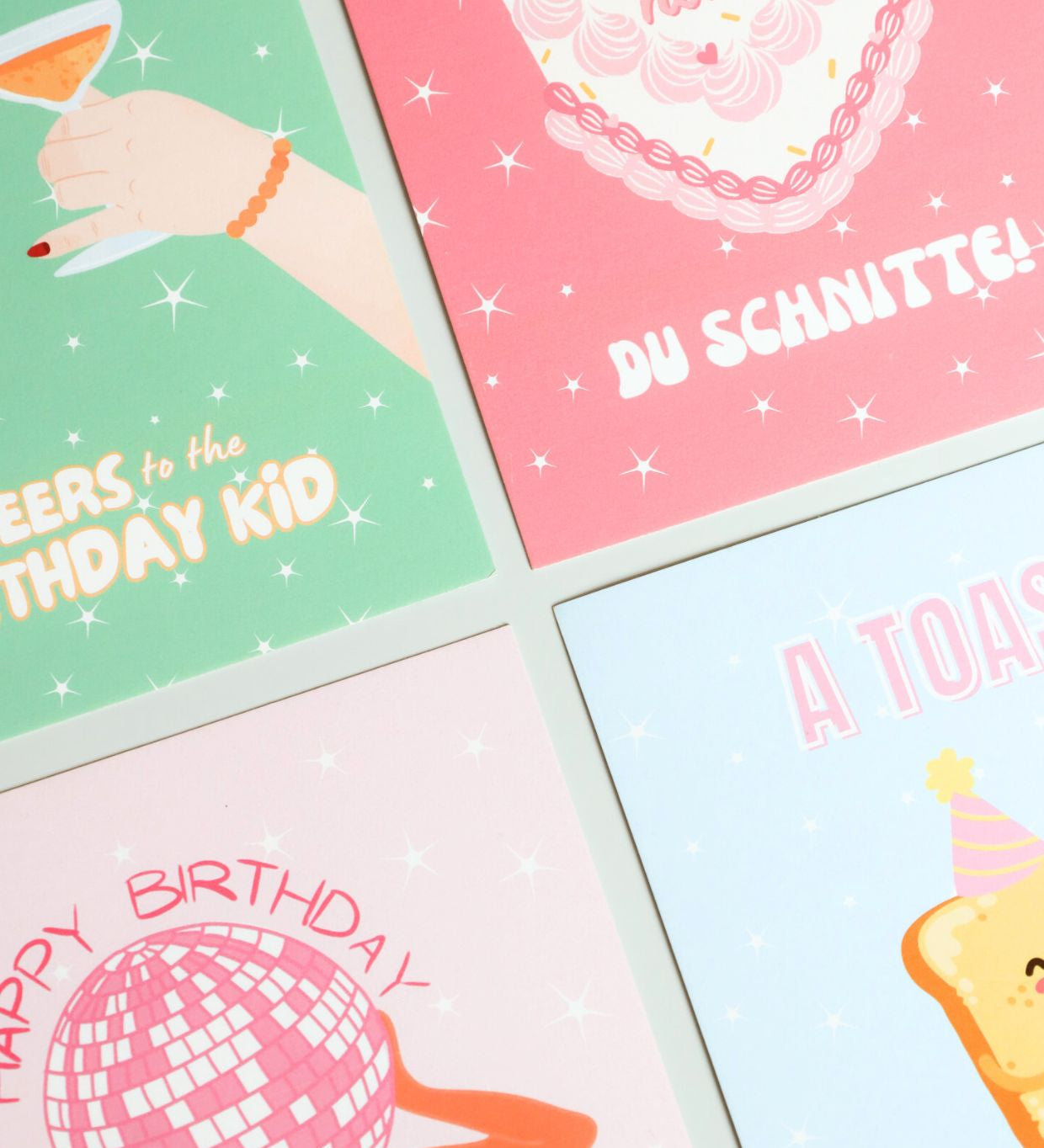 A flat lay of four colorful birthday cards with playful illustrations and text, including designs with cocktails, cake, disco ball, and toast – part of ruru. studios’ birthday card collection.