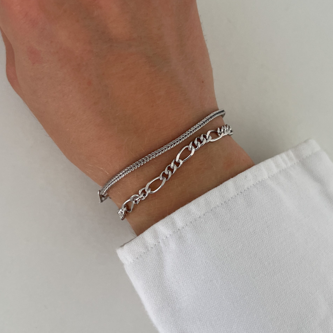 Two minimalist chain bracelets worn on wrist – one sleek wheat chain and one round braided chain, both made of 925 sterling silver.