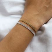 Close-up of a wrist wearing two woven mesh bracelets – one in gold and one in silver – styled against a soft white fabric background.