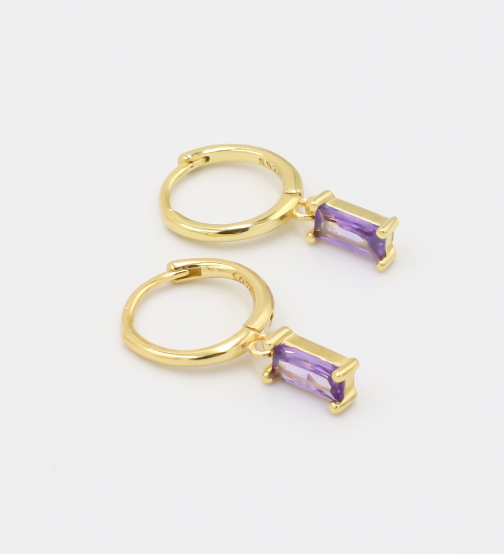 A gold-colored hoop earring with a purple gemstone attached to it, displayed against a white background.