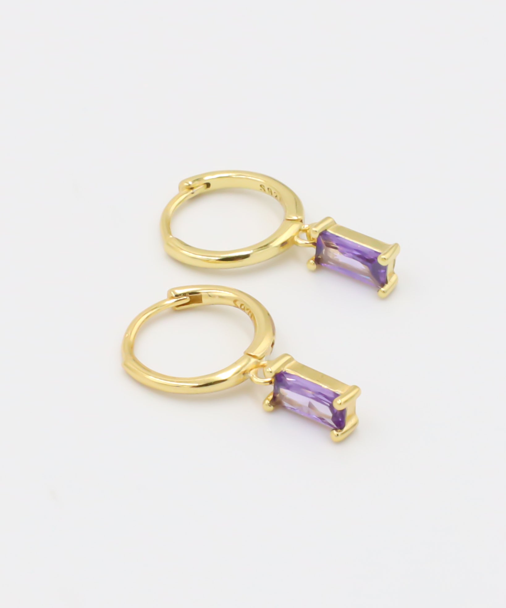 A gold-colored hoop earring with a purple gemstone attached to it, displayed against a white background.