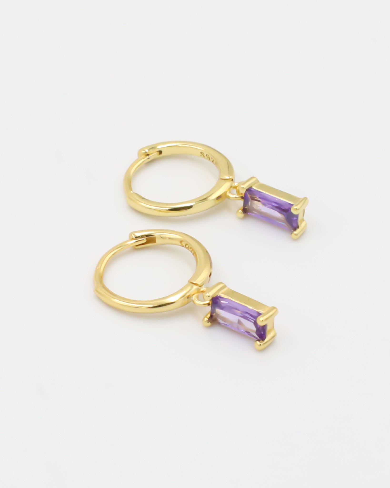 A gold-colored hoop earring with a purple gemstone attached to it, displayed against a white background.