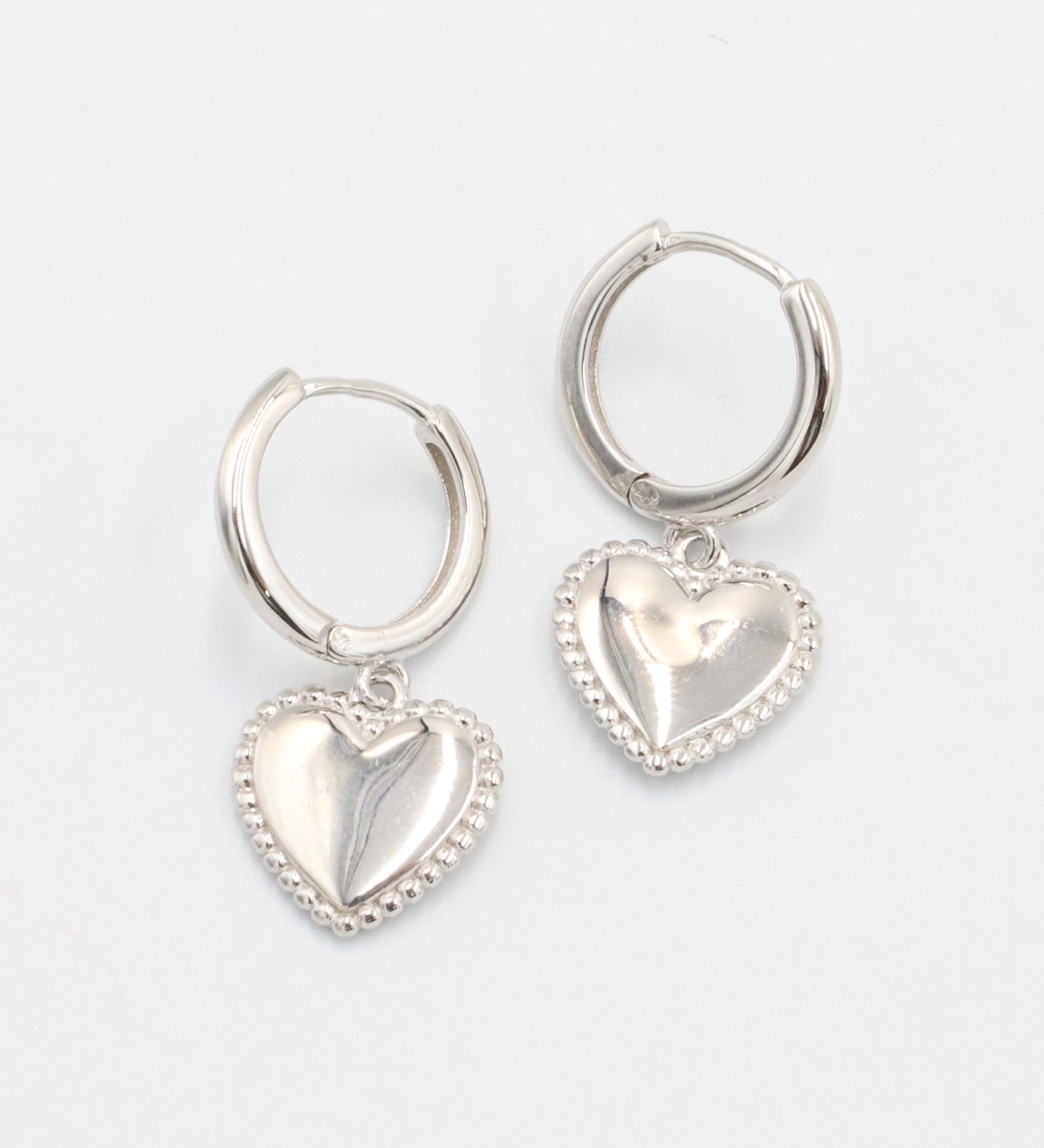 A pair of silver hoop earrings with heart-shaped pendants attached to them, displayed against a white background.