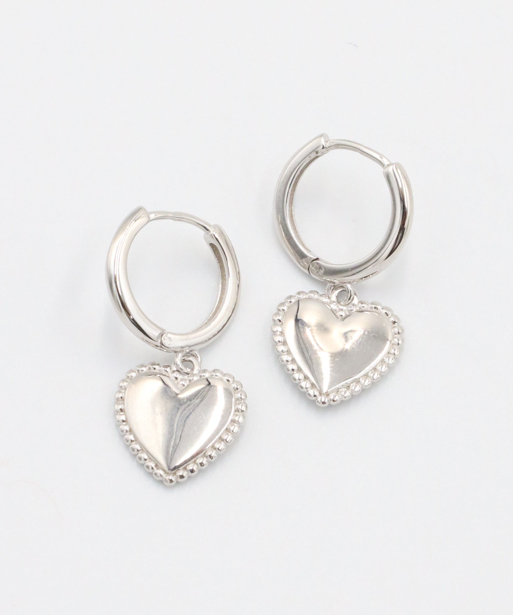 A pair of silver hoop earrings with heart-shaped pendants attached to them, displayed against a white background.