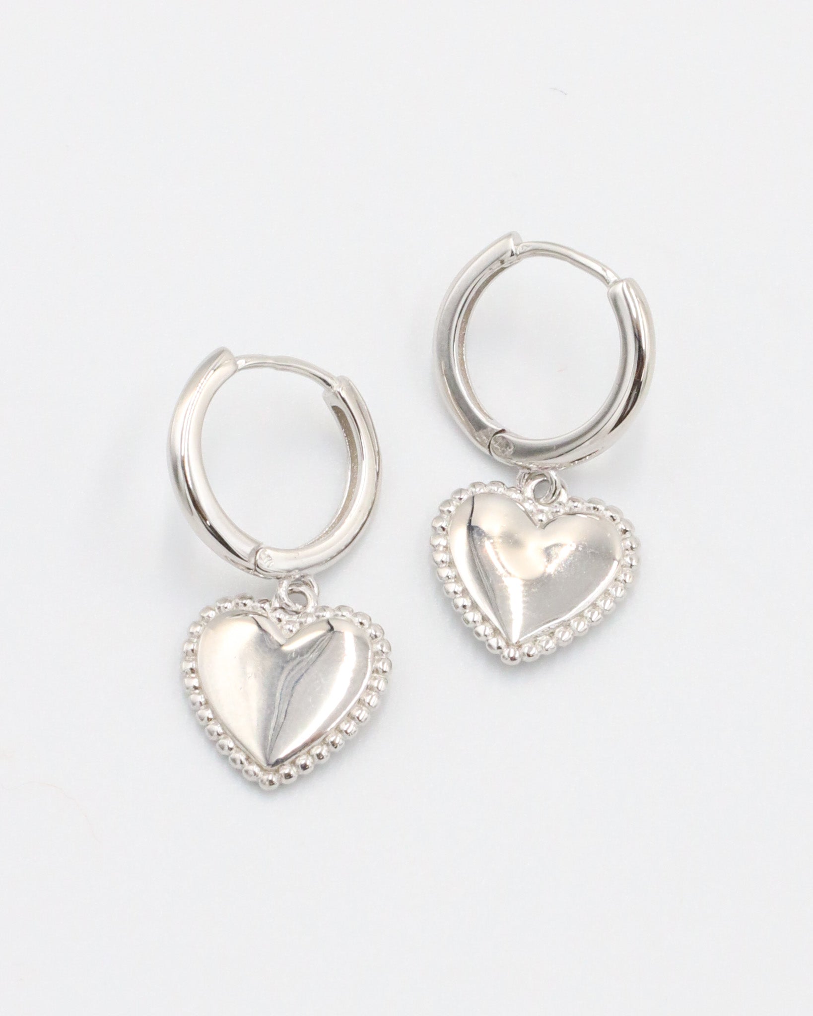 A pair of silver hoop earrings with heart-shaped pendants attached to them, displayed against a white background.