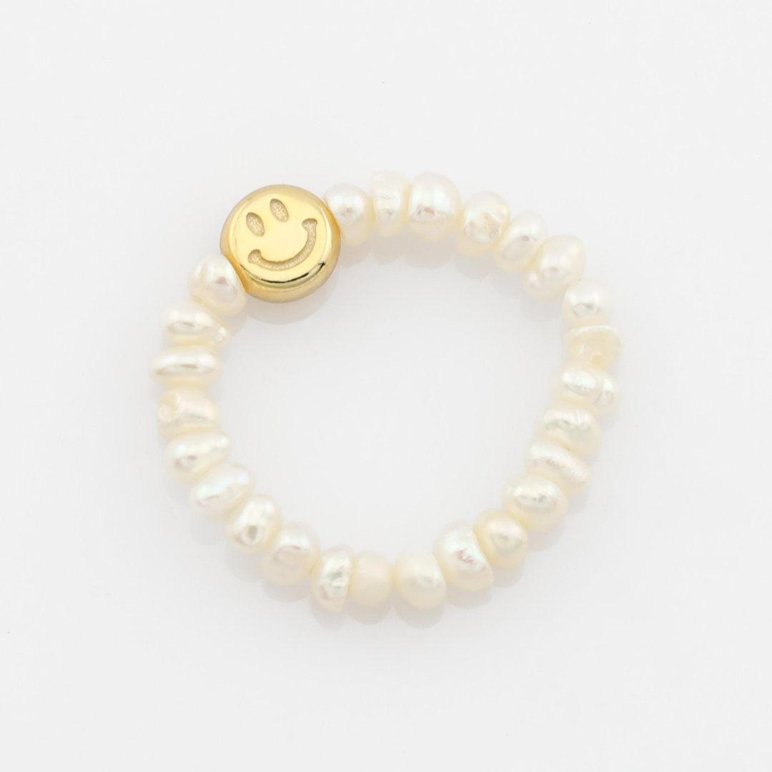 Pearl ring with a gold smiley face charm on a light gray background
