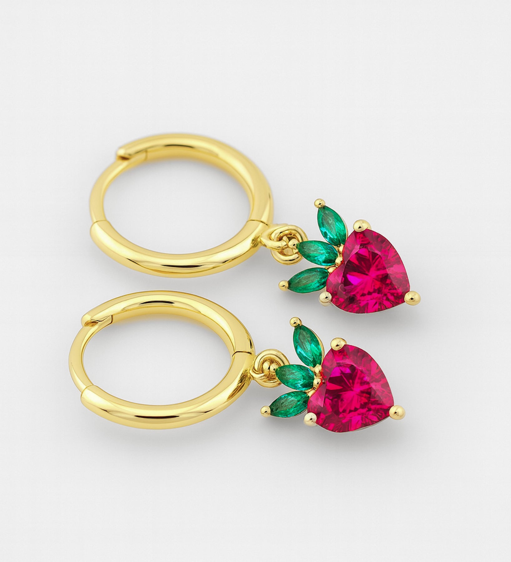 Gold hoop earrings with heart-shaped red gemstones and green leaves on a white background