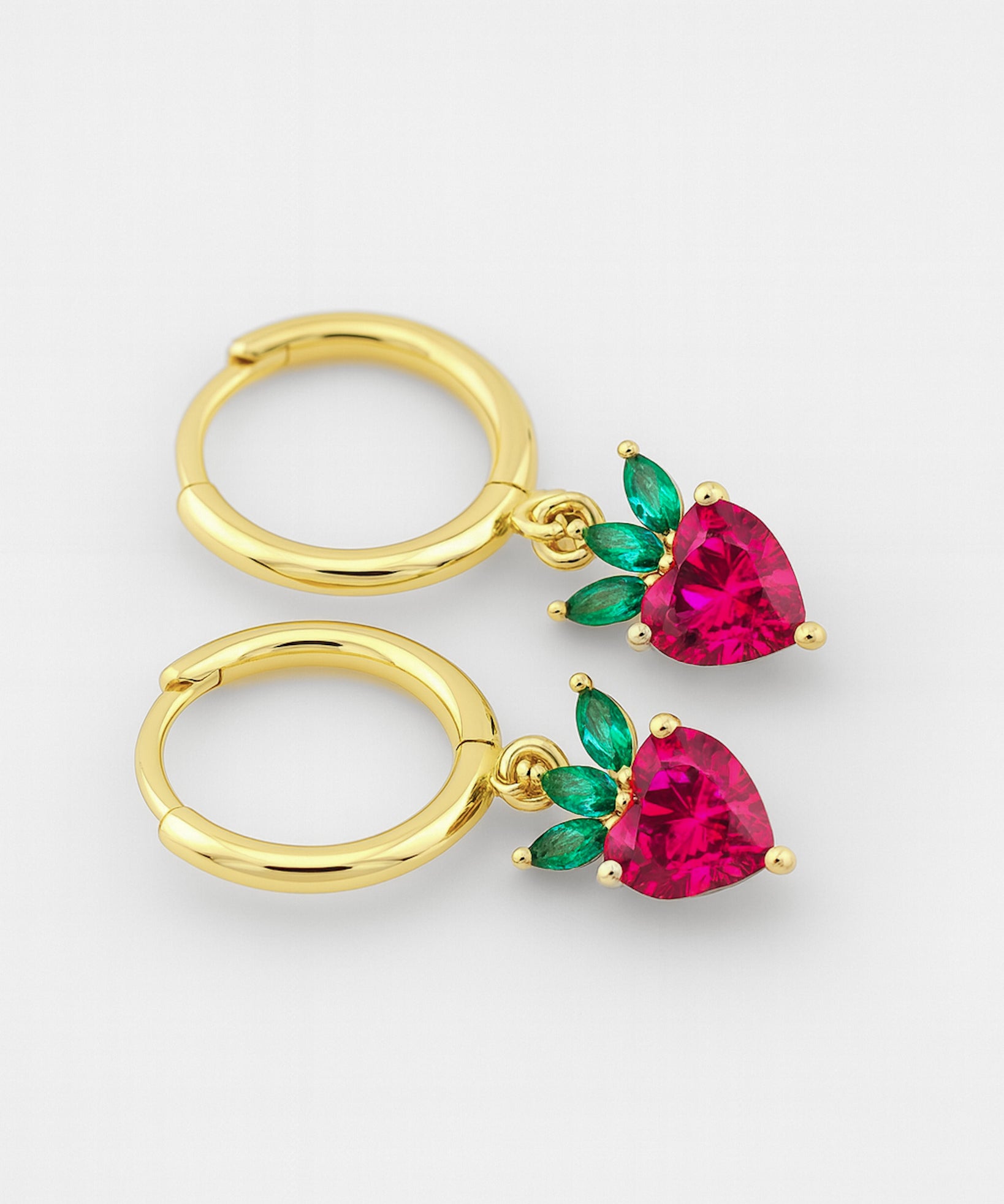 Gold hoop earrings with heart-shaped red gemstones and green leaves on a white background