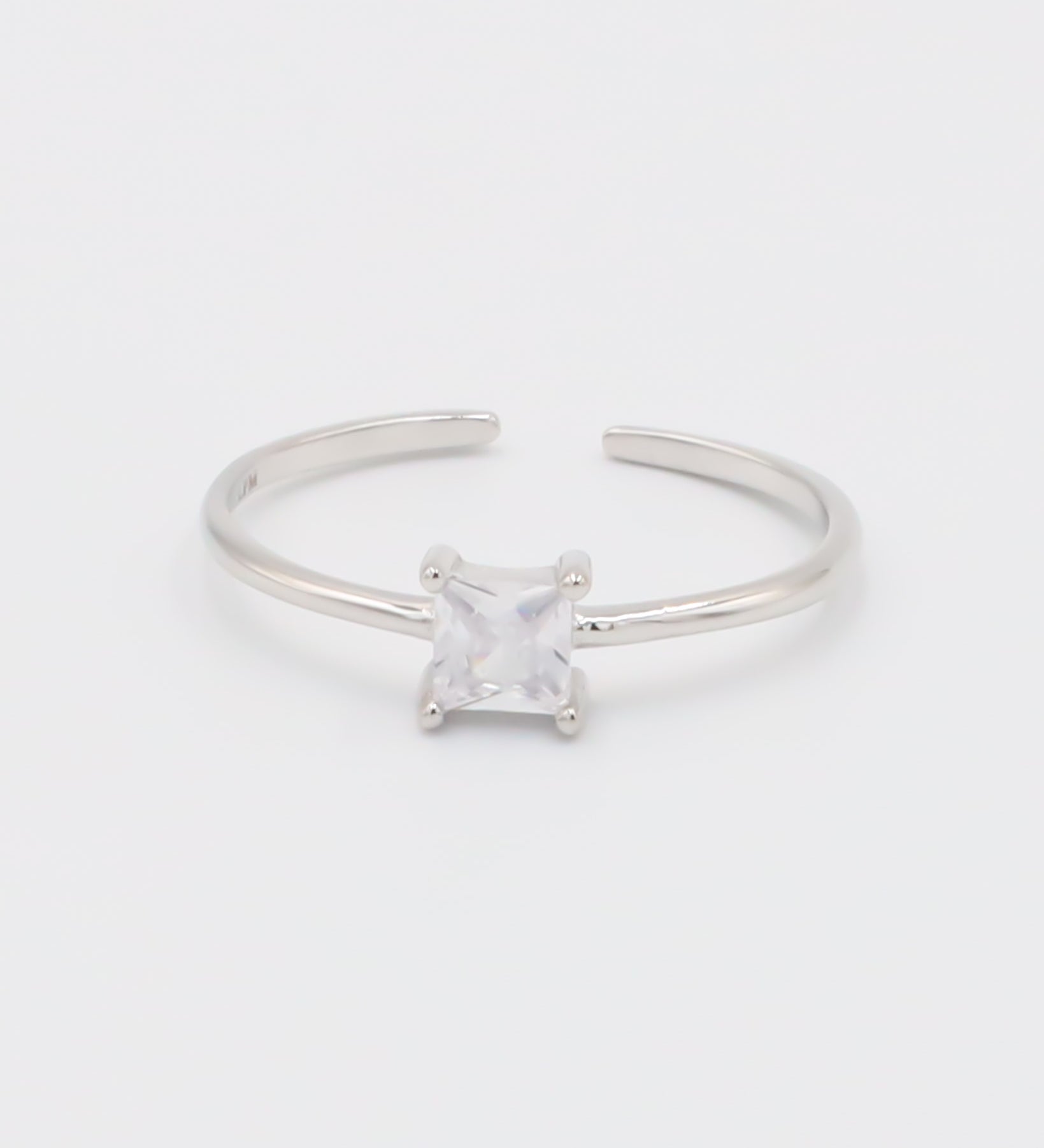A silver ring with a clear, square-cut central gemstone, surrounded by smaller clear gemstones set in a recycled 925 sterling silver band, with a textured white background.