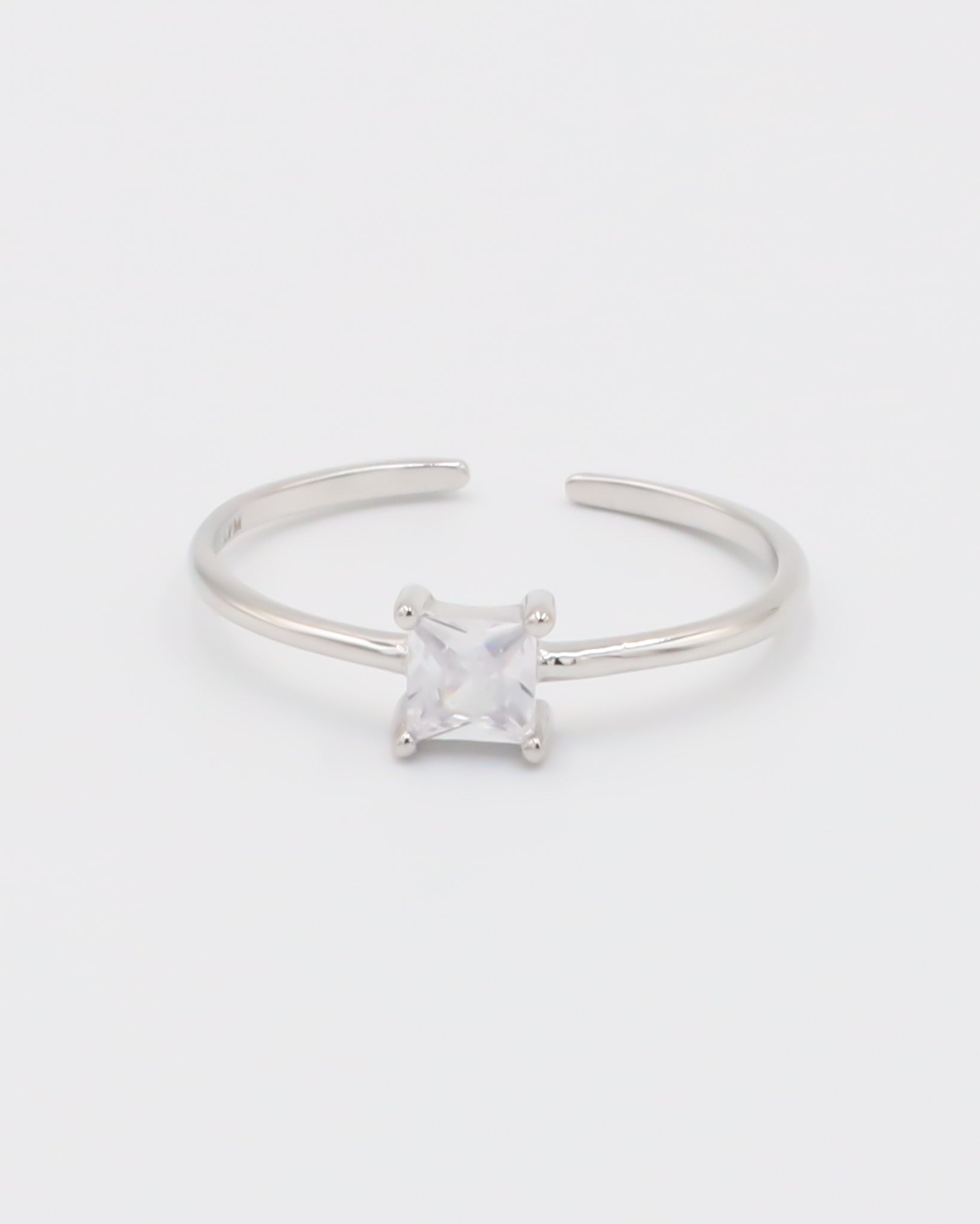 A silver ring with a clear, square-cut central gemstone, surrounded by smaller clear gemstones set in a recycled 925 sterling silver band, with a textured white background.