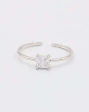 A silver ring with a clear, square-cut central gemstone, surrounded by smaller clear gemstones set in a recycled 925 sterling silver band, with a textured white background.