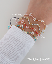 Four Roxy Bracelets in different colors – Red, Blue, Grey, and Light Pink – stacked on a wrist, with each color labeled. The bracelets feature a zigzag beaded pattern with tiny flower details.