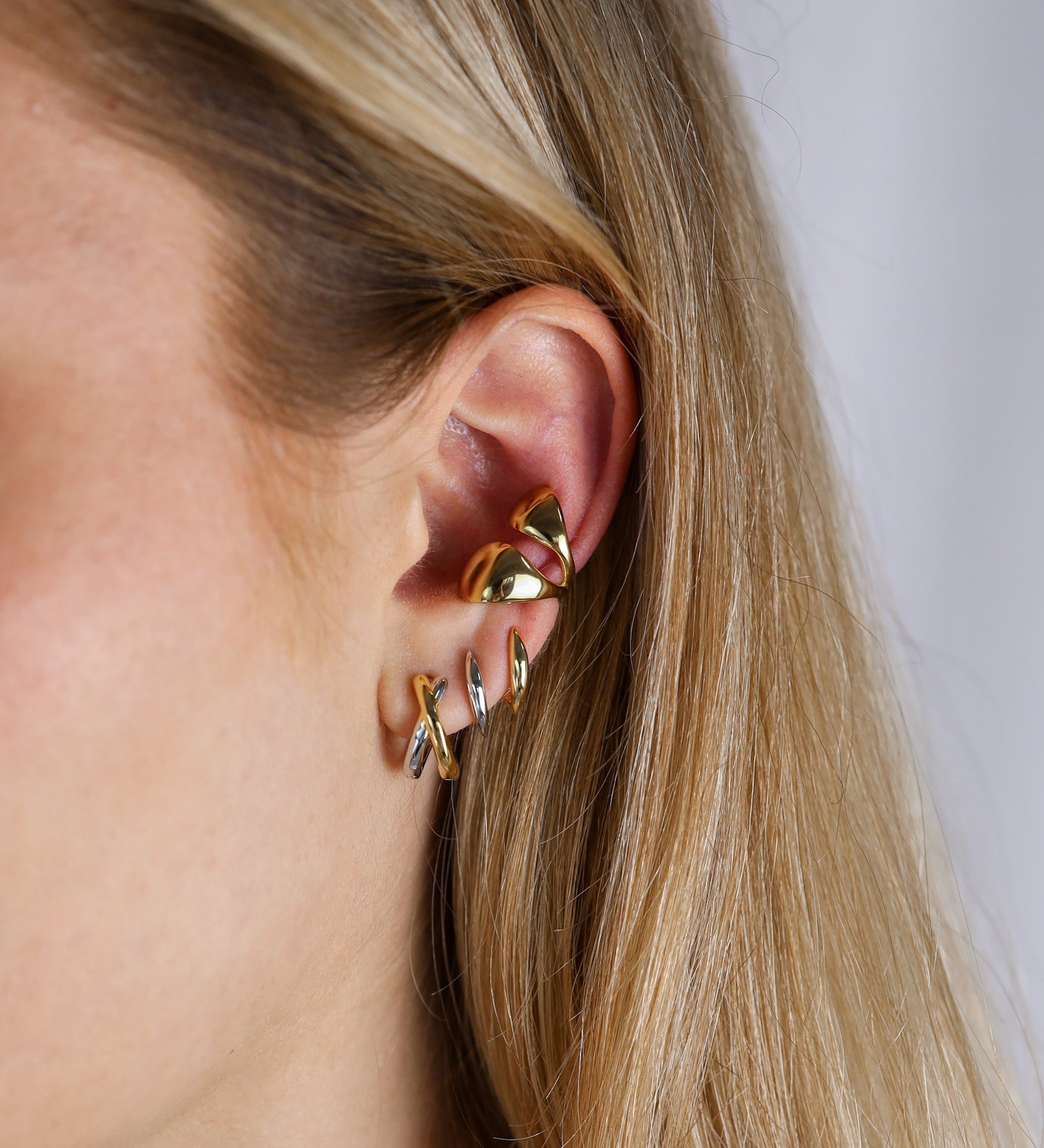 Gold earrings worn by a person with blonde hair on a neutral background