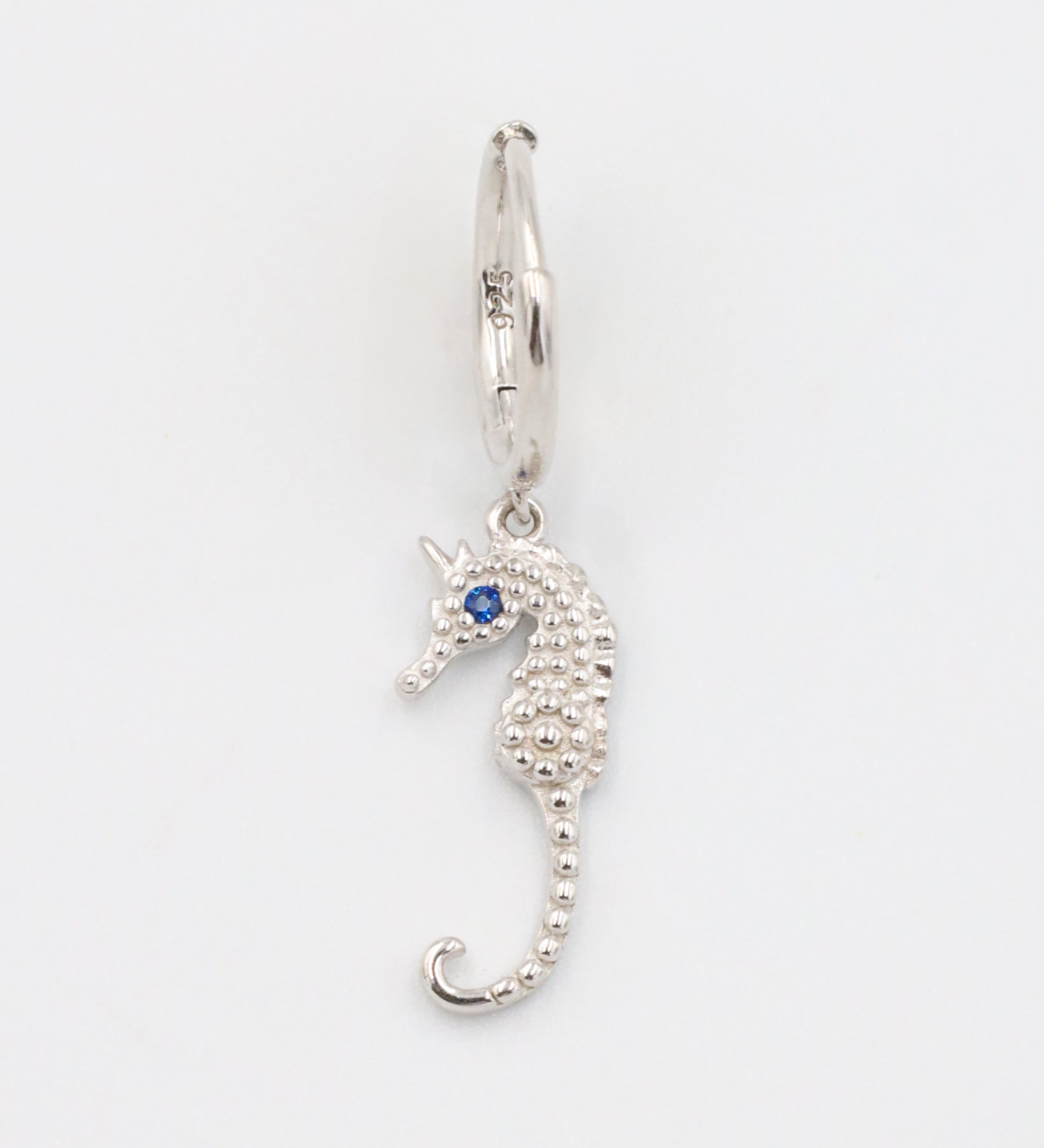 A close-up of a single silver hoop earring with a seahorse charm and a blue stone eye, displayed on a white background (Left Ear).
