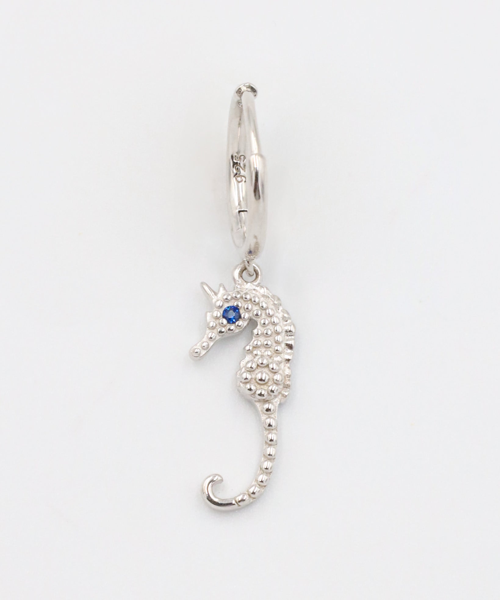 A close-up of a single silver hoop earring with a seahorse charm and a blue stone eye, displayed on a white background (Left Ear).
