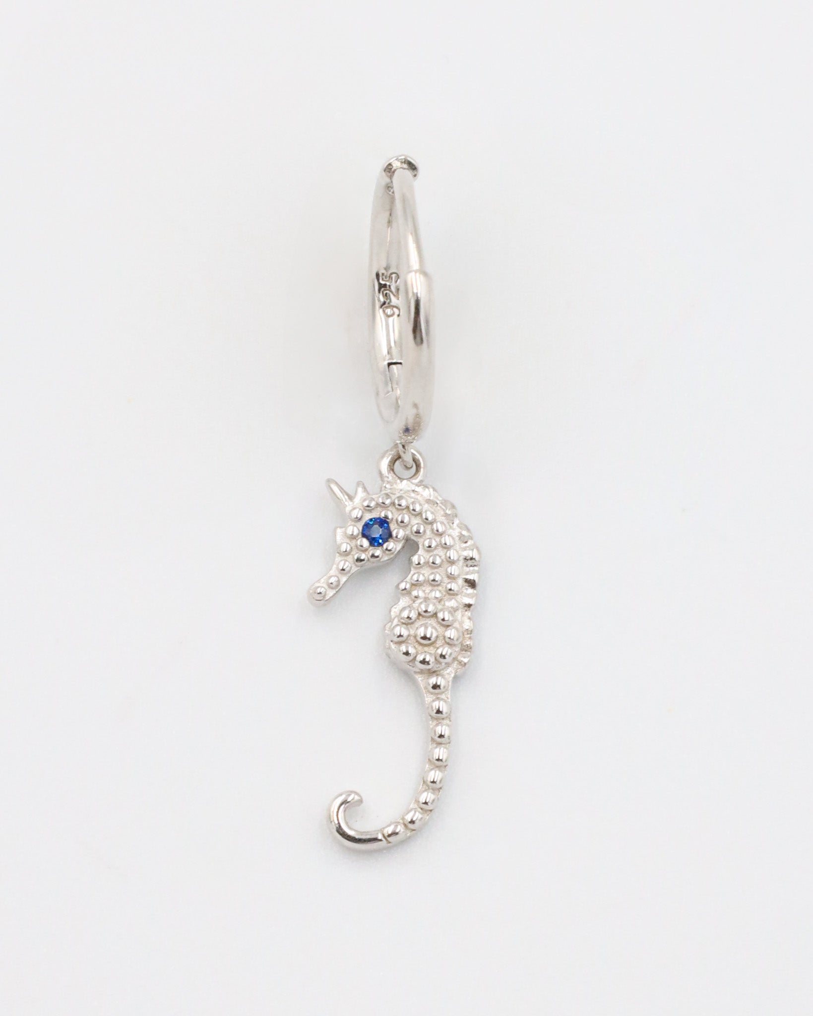 A close-up of a single silver hoop earring with a seahorse charm and a blue stone eye, displayed on a white background (Left Ear).
