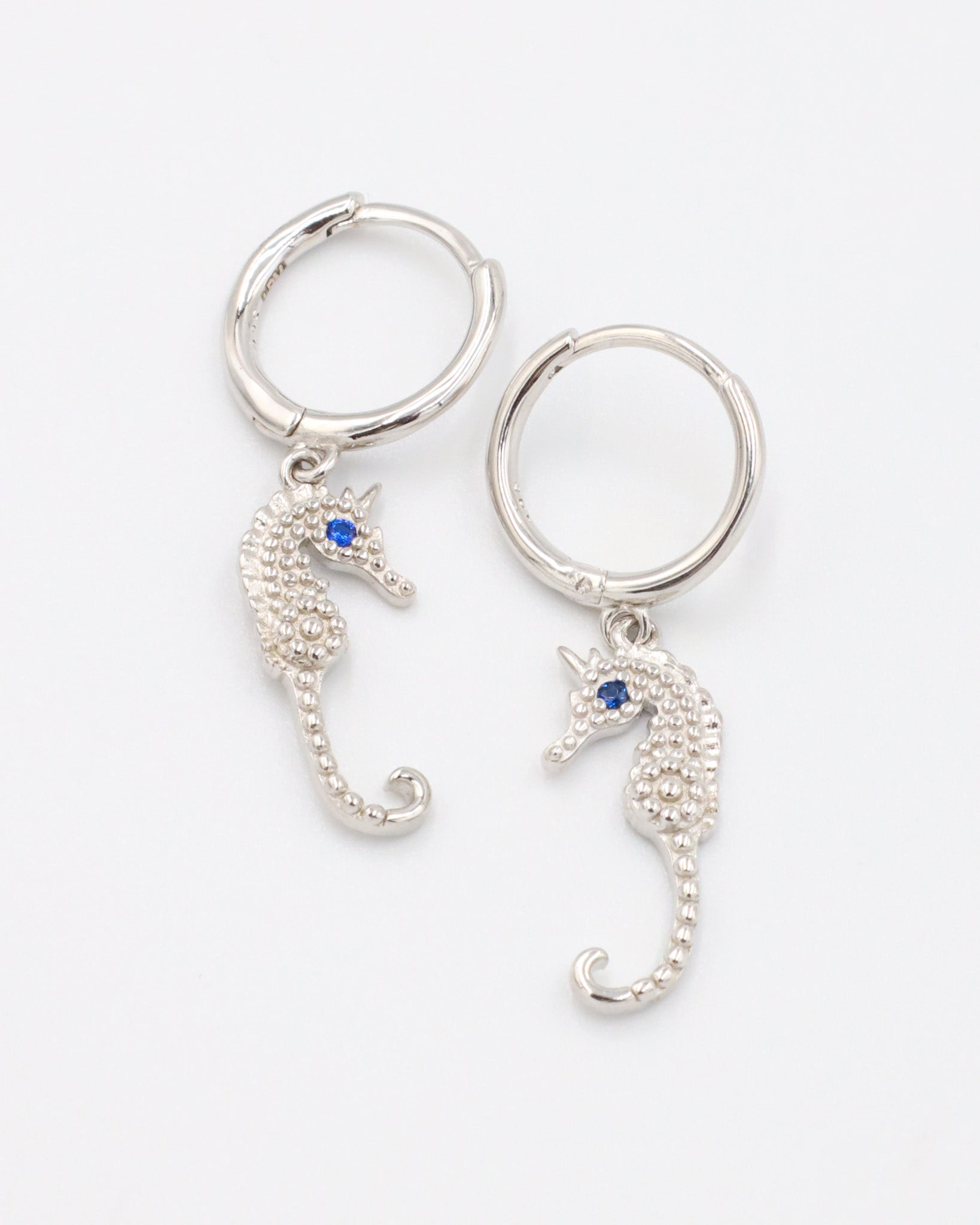 A pair of silver hoop earrings featuring detailed seahorse charms with blue stone eyes, displayed on a white background.
