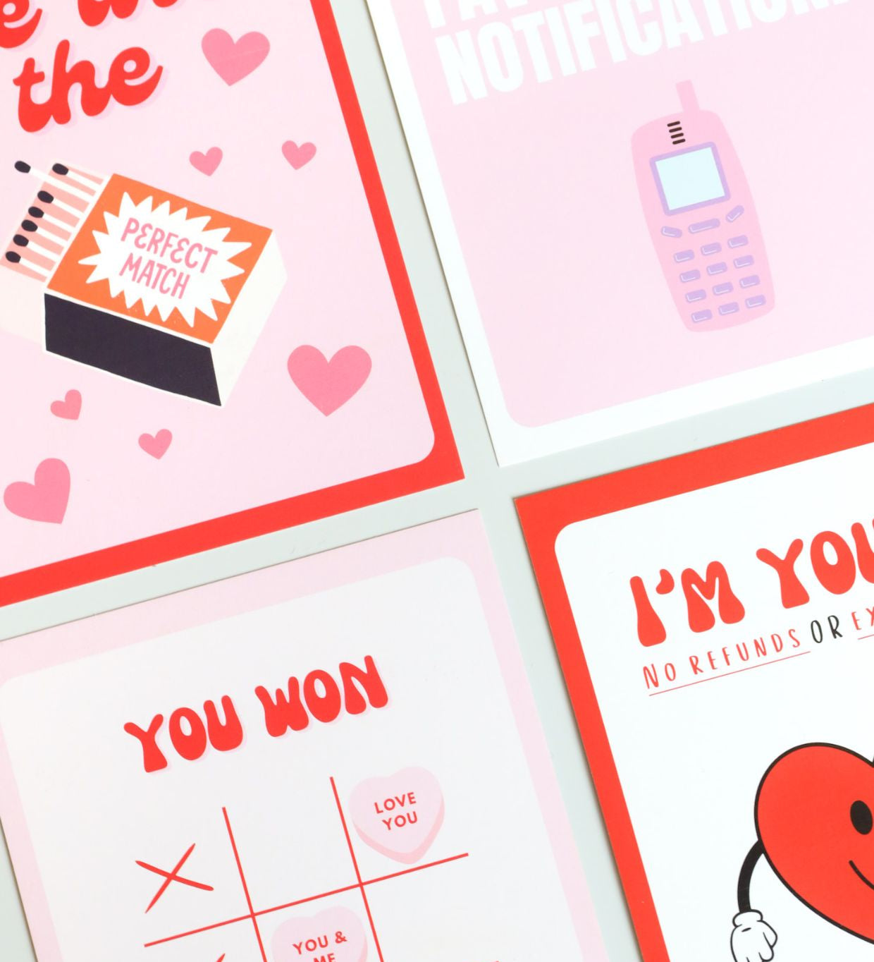 Four playful Valentine’s Day postcards in a bold, pop-art style: one with a matchbox reading “Perfect Match” surrounded by hearts, one saying “You’re my favorite notification” with a retro mobile phone, one with “You won” and a Tic-Tac-Toe layout using candy hearts, and one featuring a smiling heart with the phrase “I’m yours – no refunds.” 