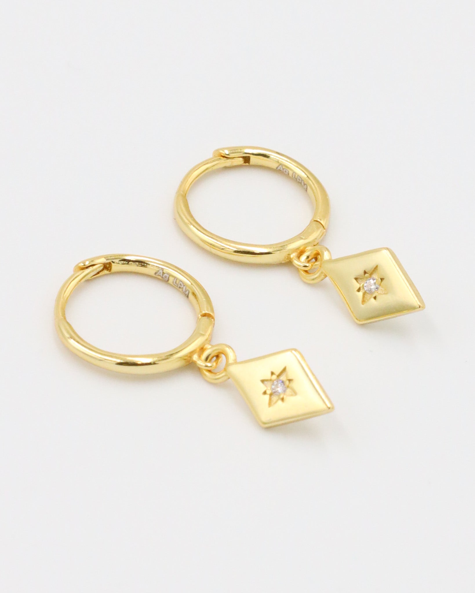 A pair of Patricia Gold earrings featuring a starburst design with a small zirconia in the center, hanging from gold hoops.
