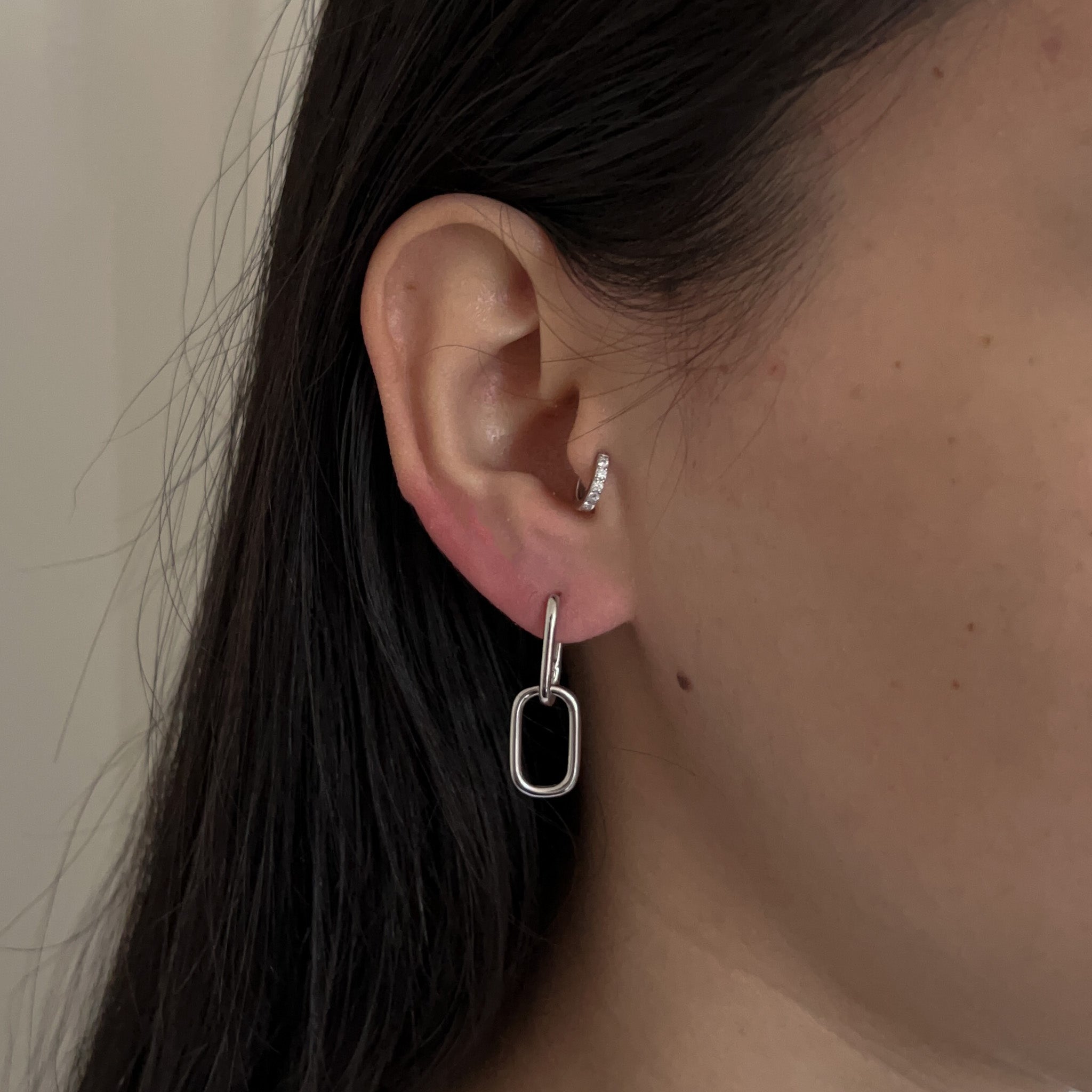 Close-up of a woman’s ear wearing two earrings: the Adelaide earring with a rectangular pendant in the lobe and the Jean Sparkly hoop in the tragus piercing, both in silver.